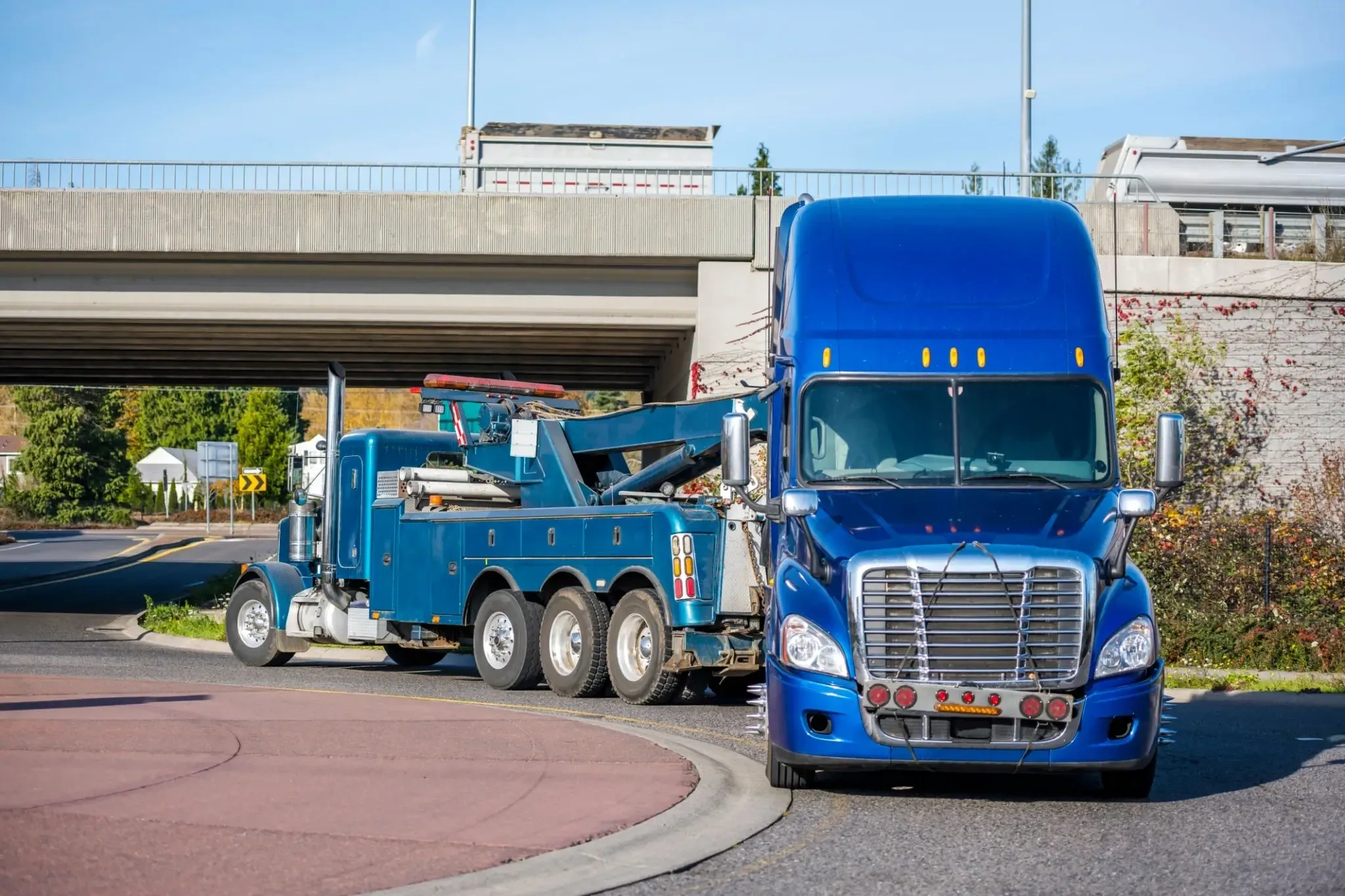 A blue semi-truck and a blue tow truck on a road near a bridge.