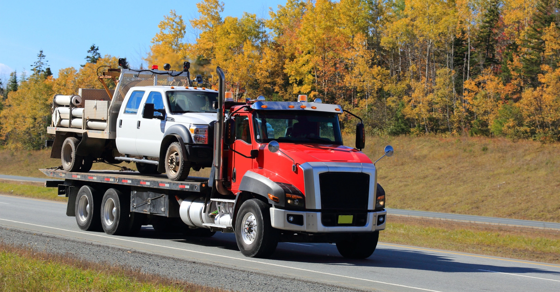 Red semi-truck transporting a white pickup truck on a highway with autumn trees in the background.